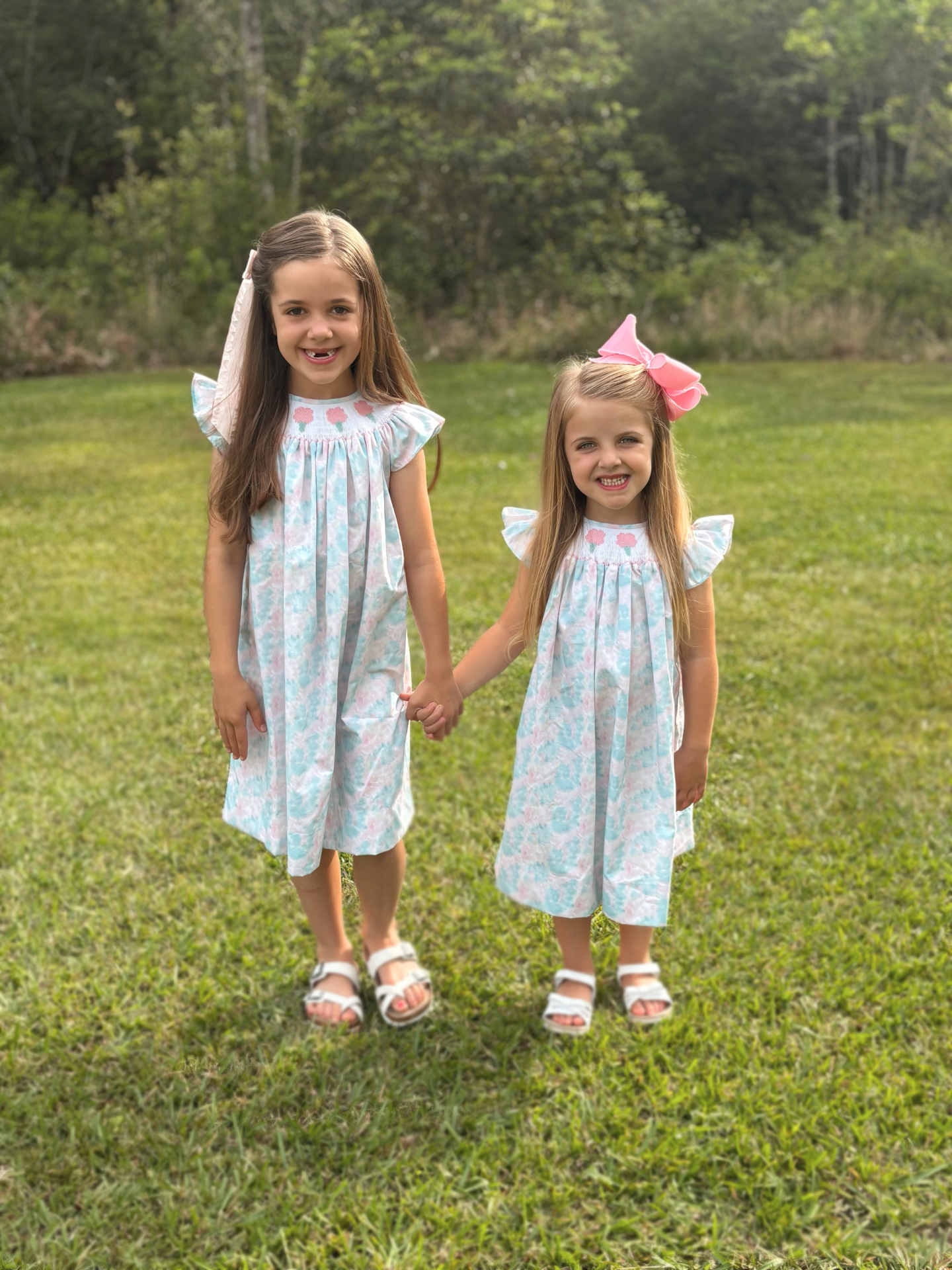 Two sisters in matching pastel smocked bishop dresses holding hands in a sunlit field